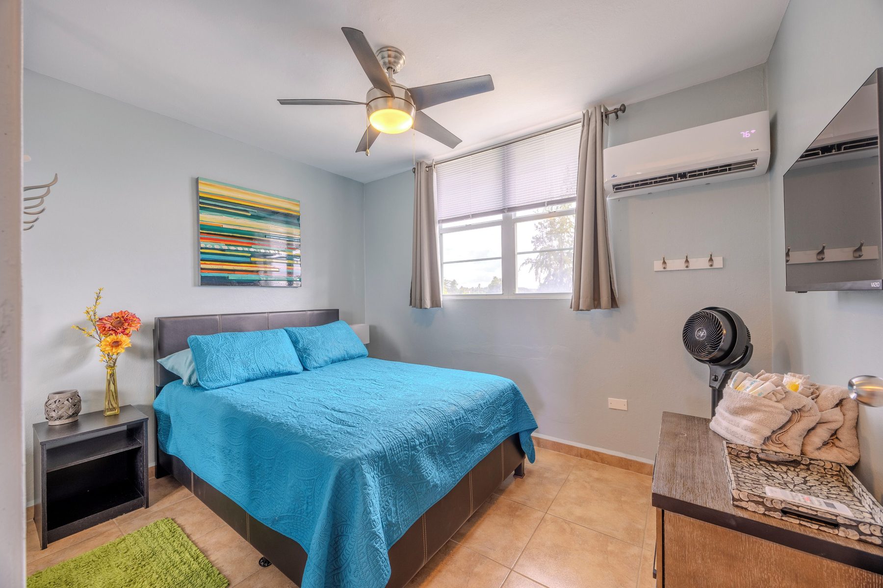 A bedroom with a queen bed, ceiling fan, ductless air conditioning, wall-mounted TV and folded towels on the dresser