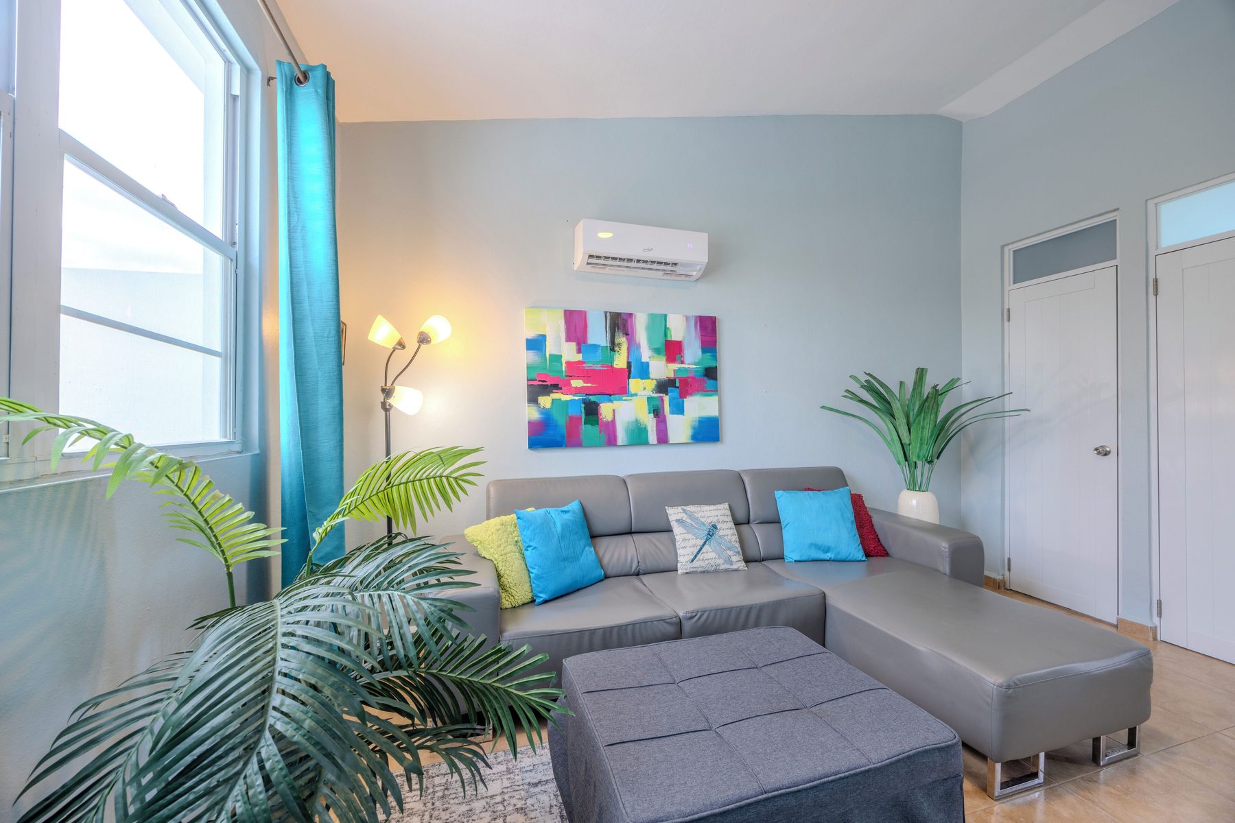 The living room with a grey sectional sofa, ottoman, floor lamp, ductless air conditioning unit and a bright window