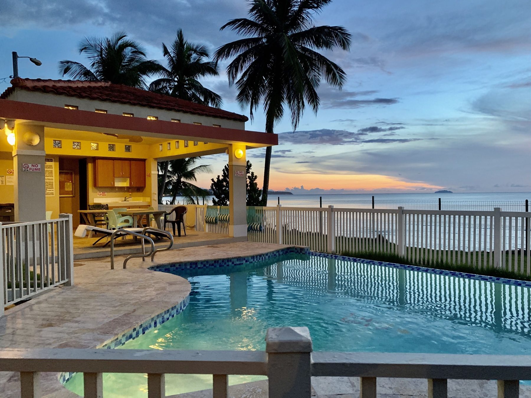 The shared pool at dusk, lit from within, with the poolside gazebo and kitchen area glowing on the left and the ocean behind the fence