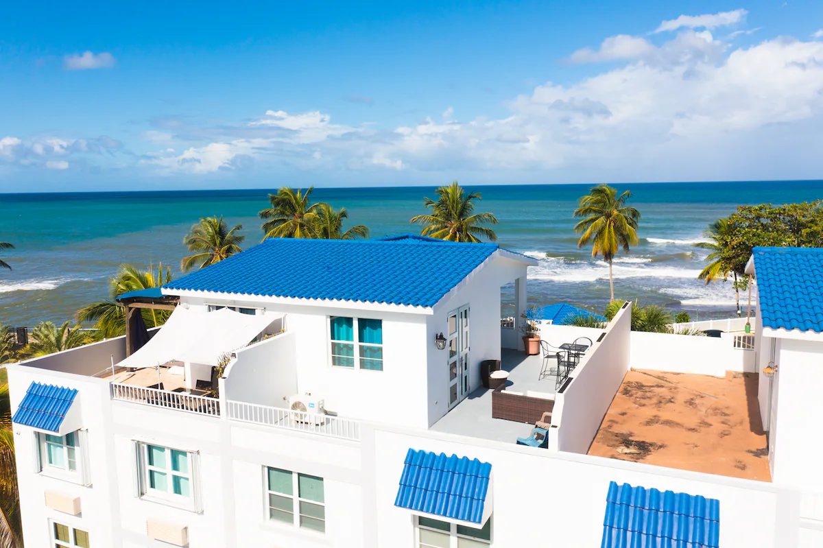 The penthouse rooftop terrace seen from above: a white unit with a blue tile roof, shaded seating and a walled terrace, with palms and the open Atlantic directly behind it