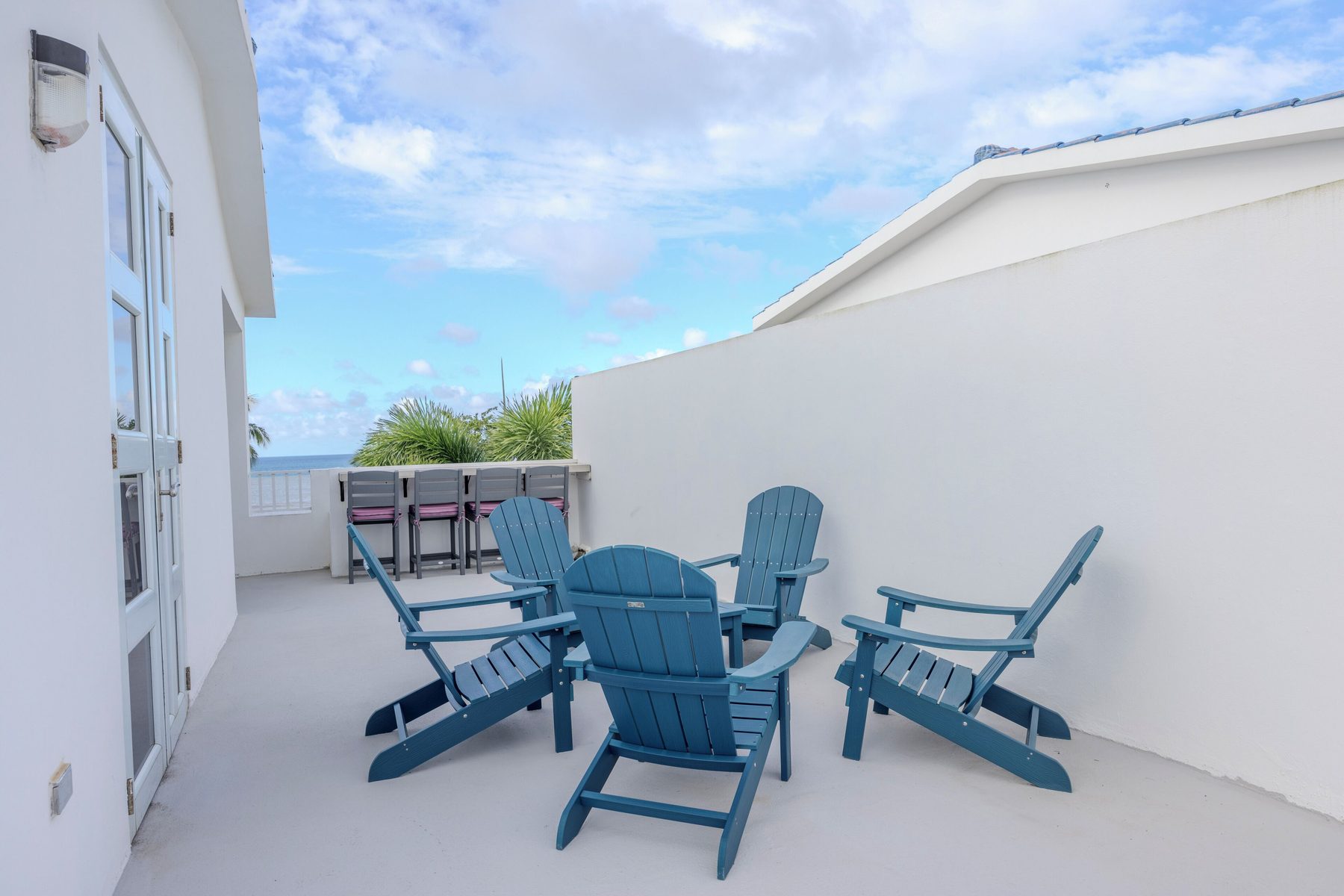Four blue Adirondack chairs arranged on the white rooftop terrace, with the bar stools and the sea visible past the wall